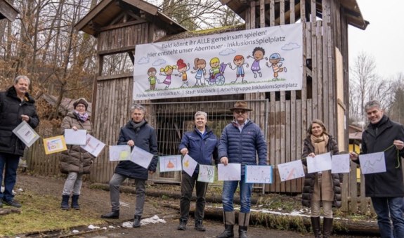 Die Mitglieder der Arbeitsgruppe InklusionsSpielplatz stehen nebeneinander vor dem Eingang des stillgelegten Spielplatzes mit gemalten Bildern von Kindern in der Hand, © Arbeitsgruppe InklusionsSpielplatz